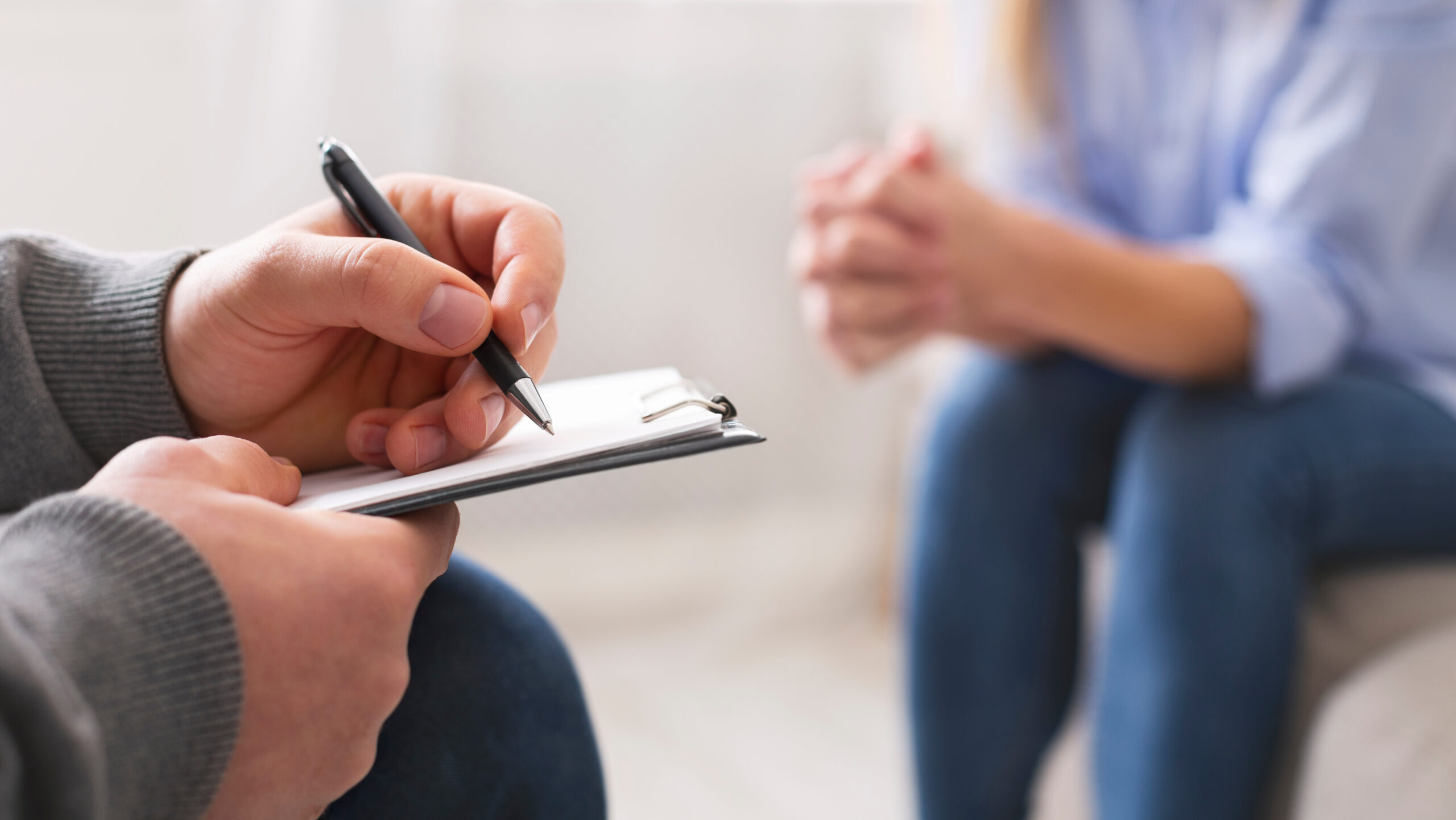 Close-up of a counseling session showing a person holding a clipboard and pen, taking notes, while another person sits across with hands clasped.